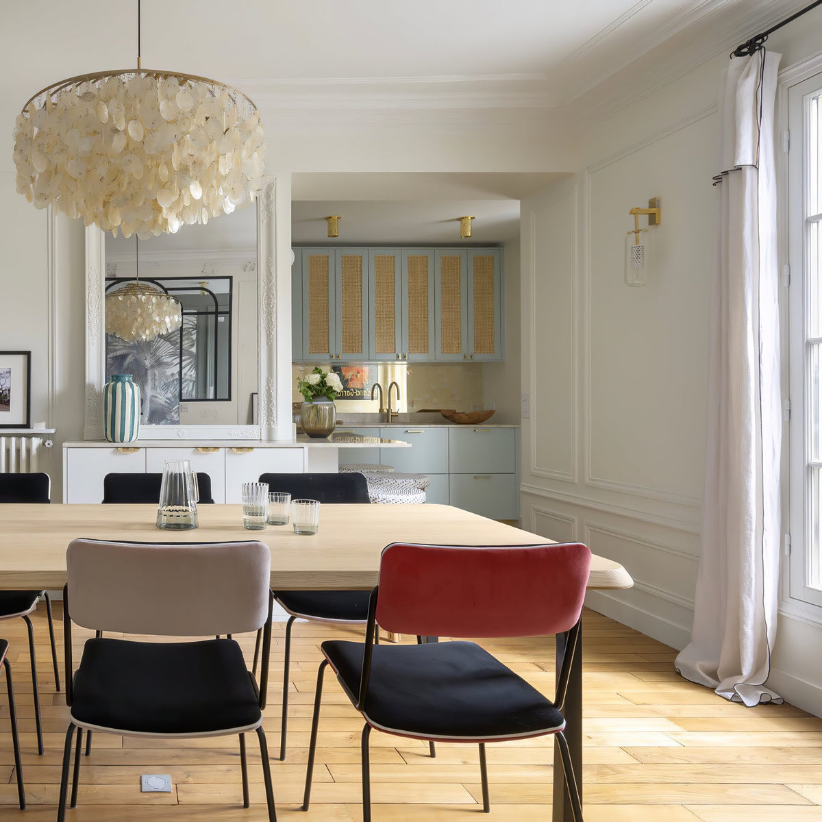 Clérachic shell chandelier at the top corner of a bright dining room with a pale wood table and red and grey chairs.