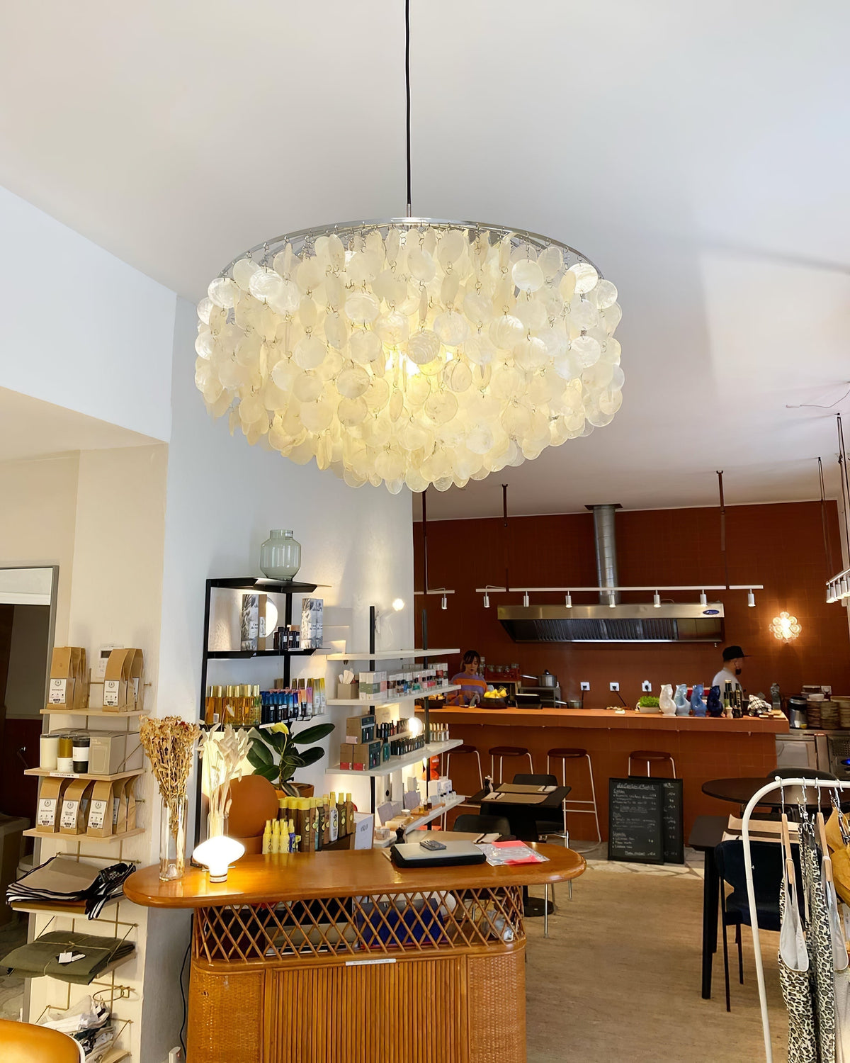 Lit Clérachic shell chandelier hung in a shop-café with a rust-red wall, a wicker counter and shelves of goods.