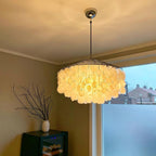 Lit Clérachic shell chandelier with a chrome ceiling cap, glowing warm yellow in a corner by a frosted window.