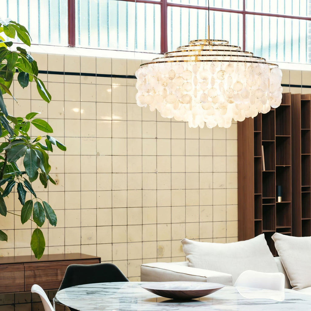 Gold-ringed Clérachic shell chandelier in a loft-style room with a tiled wall, tall windows and a marble table.