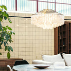 Gold-ringed Clérachic shell chandelier in a loft-style room with a tiled wall, tall windows and a marble table.