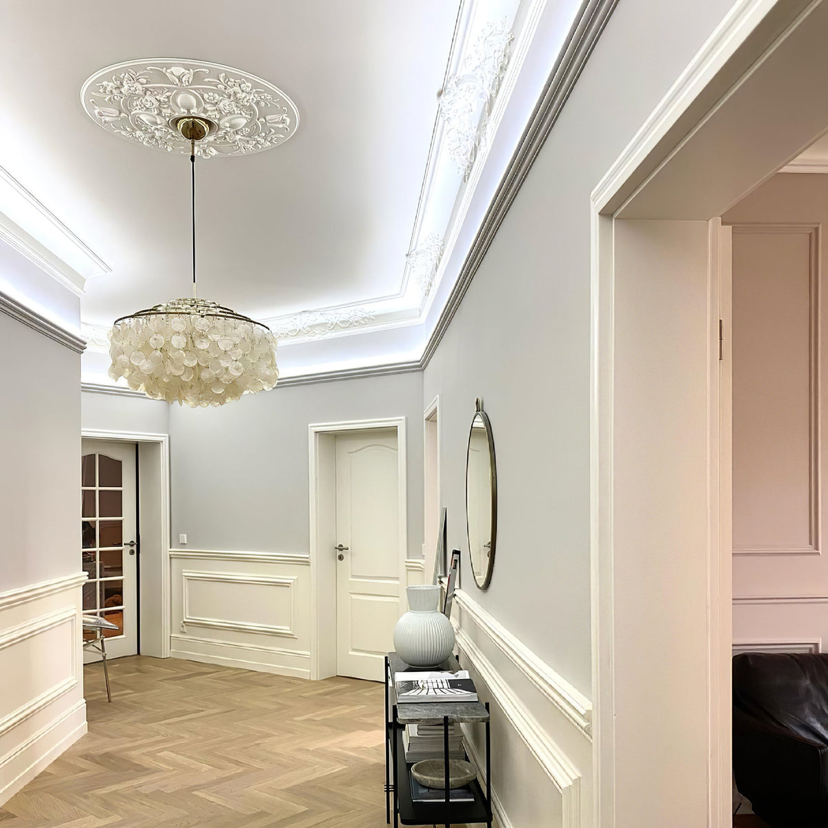 Clérachic shell chandelier hung from a carved ceiling rose in a pale hallway with white panelling and a herringbone floor.