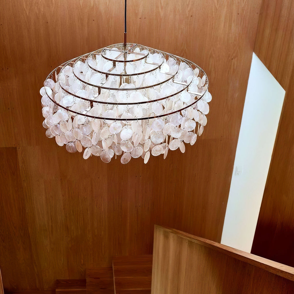 Looking up at a gold-ringed Clérachic shell chandelier above an oak-panelled stairwell, its discs lit from inside.