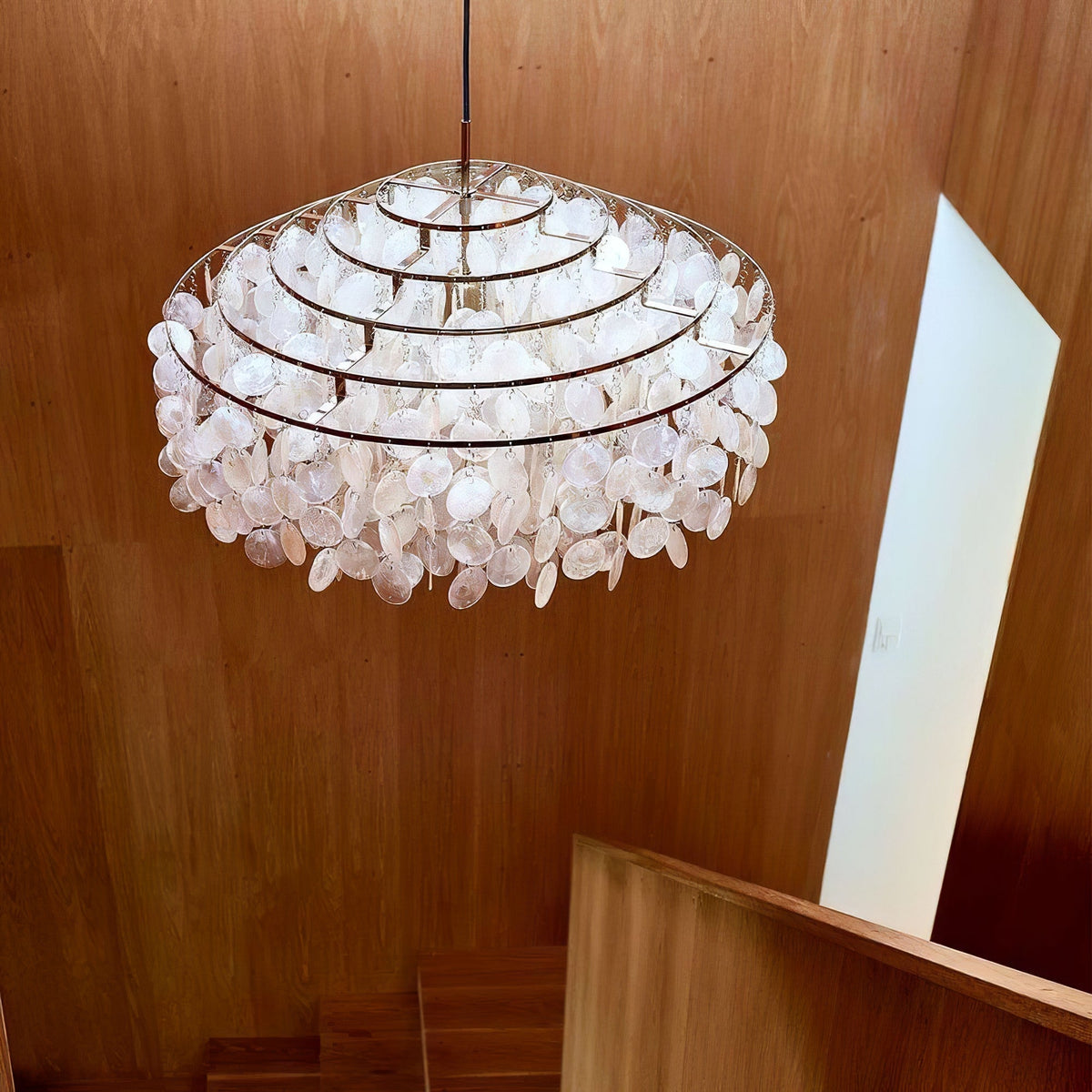 Looking up at a gold-ringed Clérachic shell chandelier above an oak-panelled stairwell, its discs lit from inside.