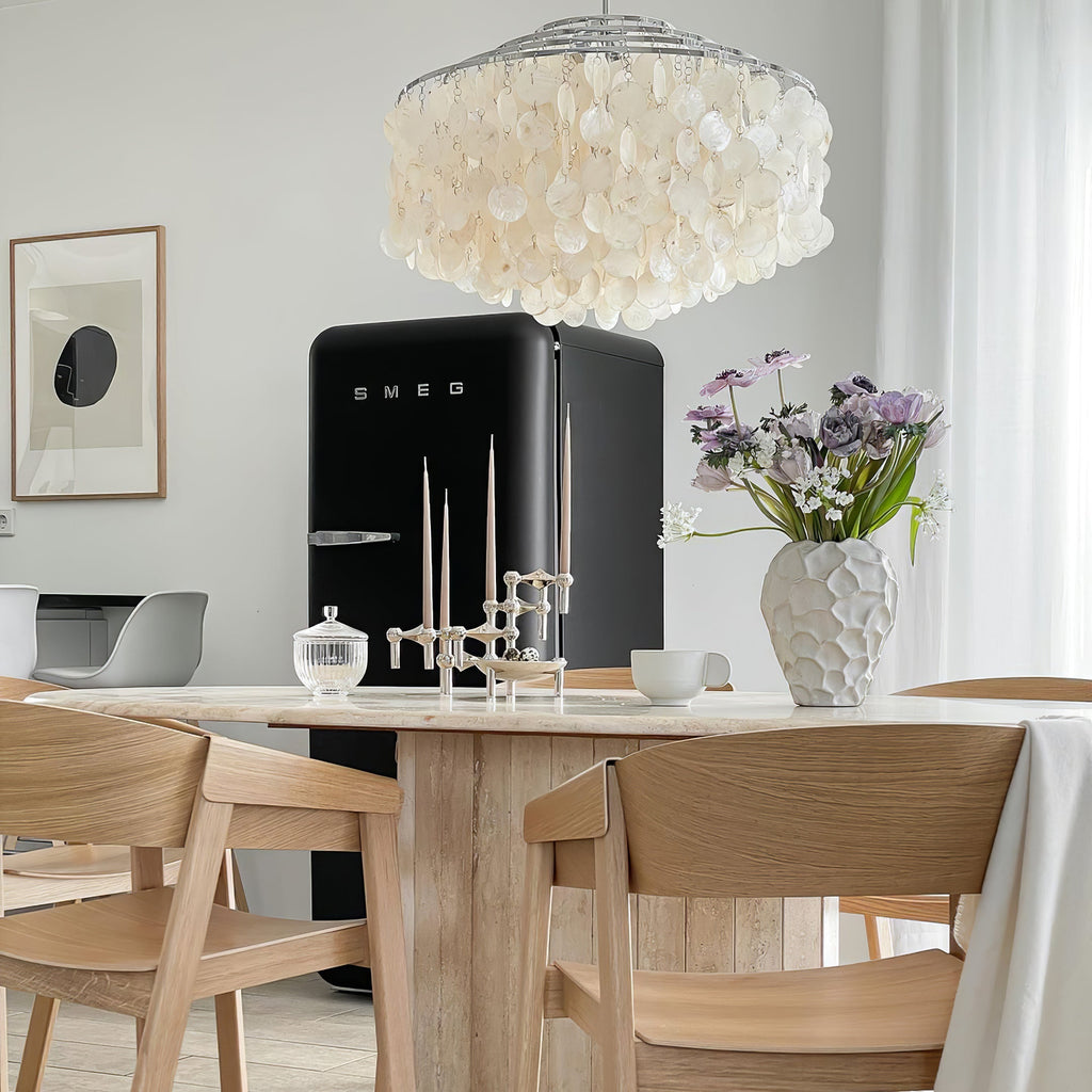 Clérachic shell chandelier above a black retro fridge, a travertine table and light oak chairs, with a vase of lilac flowers.