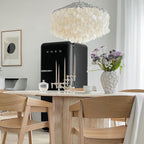 Clérachic shell chandelier above a black retro fridge, a travertine table and light oak chairs, with a vase of lilac flowers.