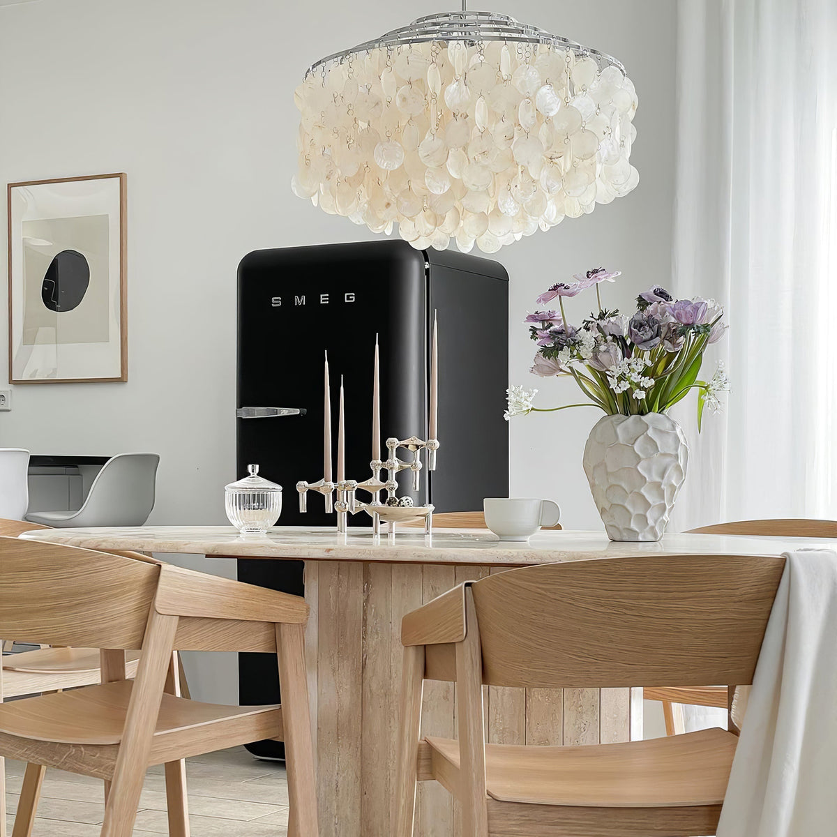 Clérachic shell chandelier above a black retro fridge, a travertine table and light oak chairs, with a vase of lilac flowers.