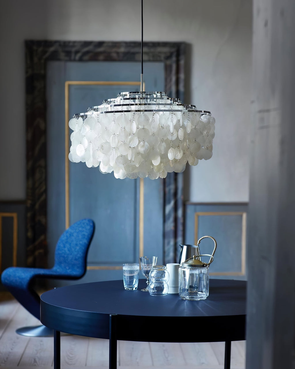 Chrome-ringed Clérachic shell chandelier against a blue-grey wall with a navy table, glassware and a blue chair below.