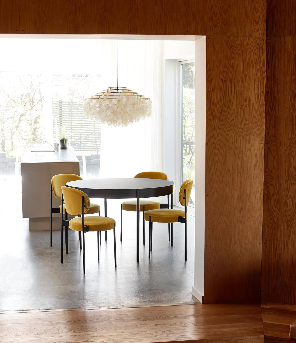 Clérachic shell chandelier seen through an oak-framed doorway, above a round table with mustard velvet chairs.