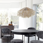 Gold-ringed Clérachic shell chandelier above a round black table and grey stools, by large windows onto a garden.