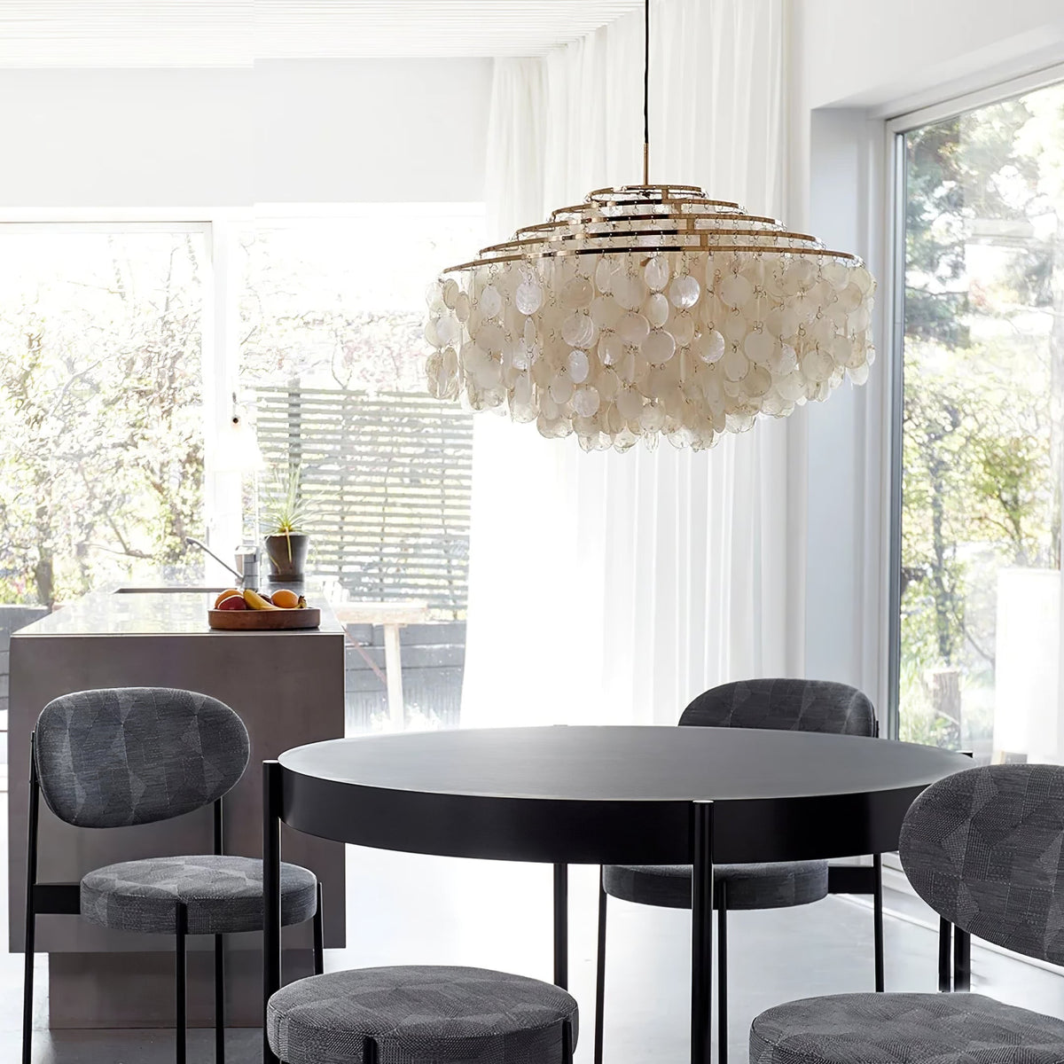 Gold-ringed Clérachic shell chandelier above a round black table and grey stools, by large windows onto a garden.