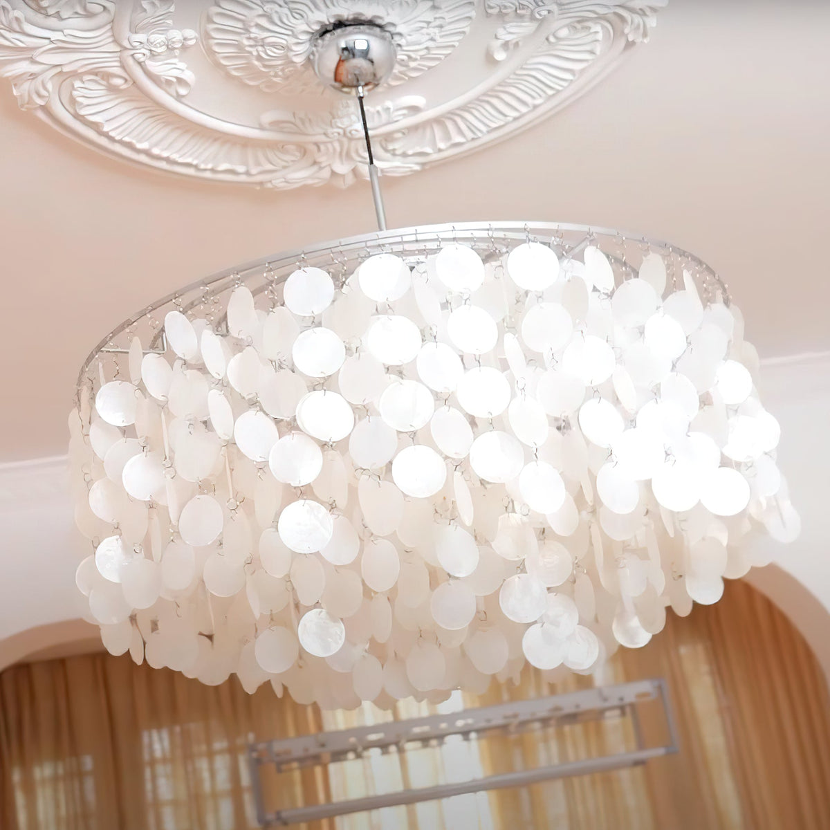 Looking up at a Clérachic shell chandelier with a chrome cap, below an ornate white ceiling rose and a warm pink wall.