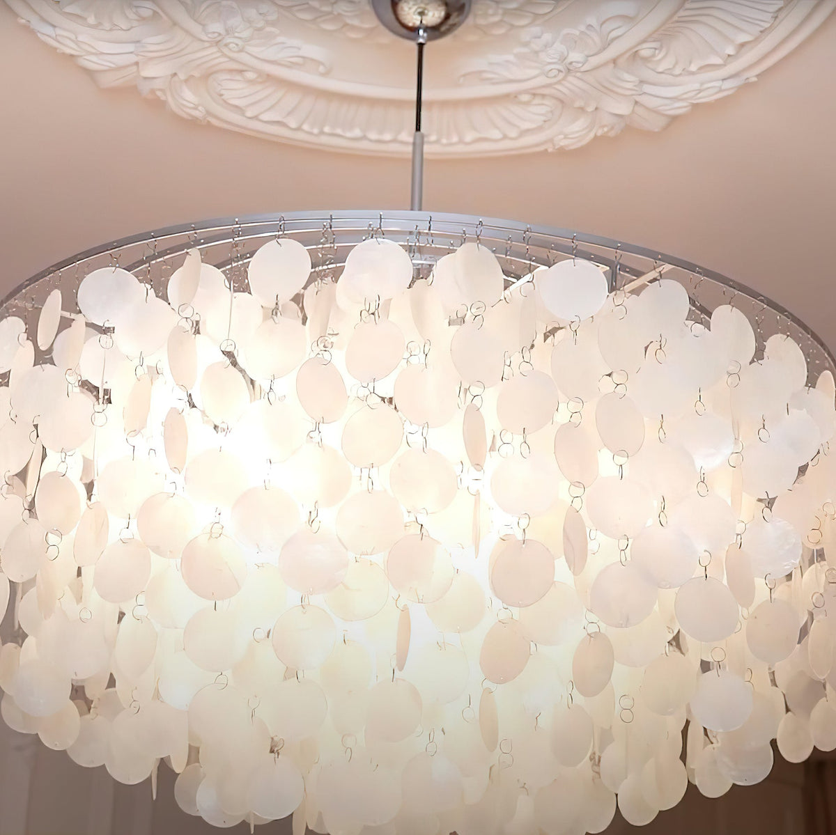 Close view of the lit shell discs inside a wide Clérachic chandelier, below a chrome cap and a plasterwork ceiling.