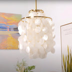 Slim gold-ringed Clérachic shell chandelier hung in front of a sunset landscape painting and a gold-framed print.