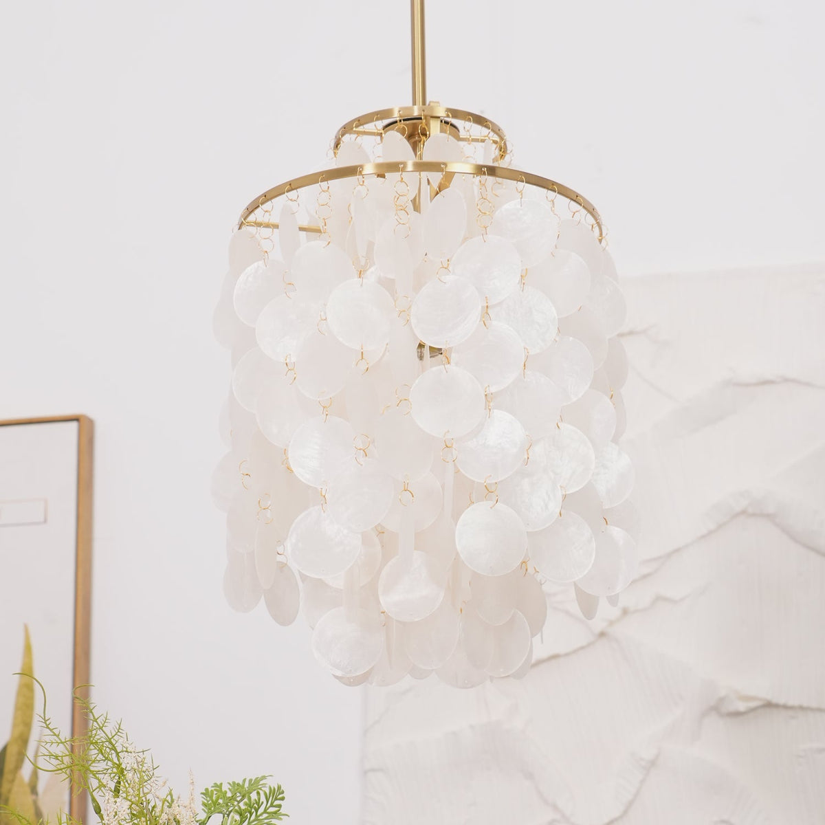 Close view of a gold-ringed Clérachic shell chandelier, white discs on small links, against a white textured wall.