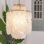 Close view of a lit gold-ringed Clérachic shell chandelier, with banana leaves and an arched window behind it.