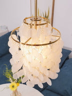Looking up into a lit gold-ringed Clérachic shell chandelier with its spoke frame visible, a blue sofa behind it.