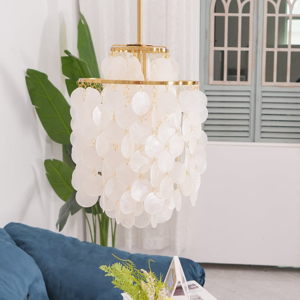 Unlit gold-ringed Clérachic shell chandelier beside banana leaves and arched shuttered windows, with a blue sofa below.