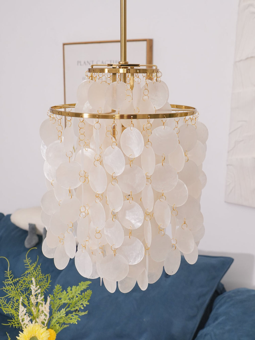 Close view of a gold-ringed Clérachic shell chandelier, discs hanging in two tiers beside a framed print and a white wall.