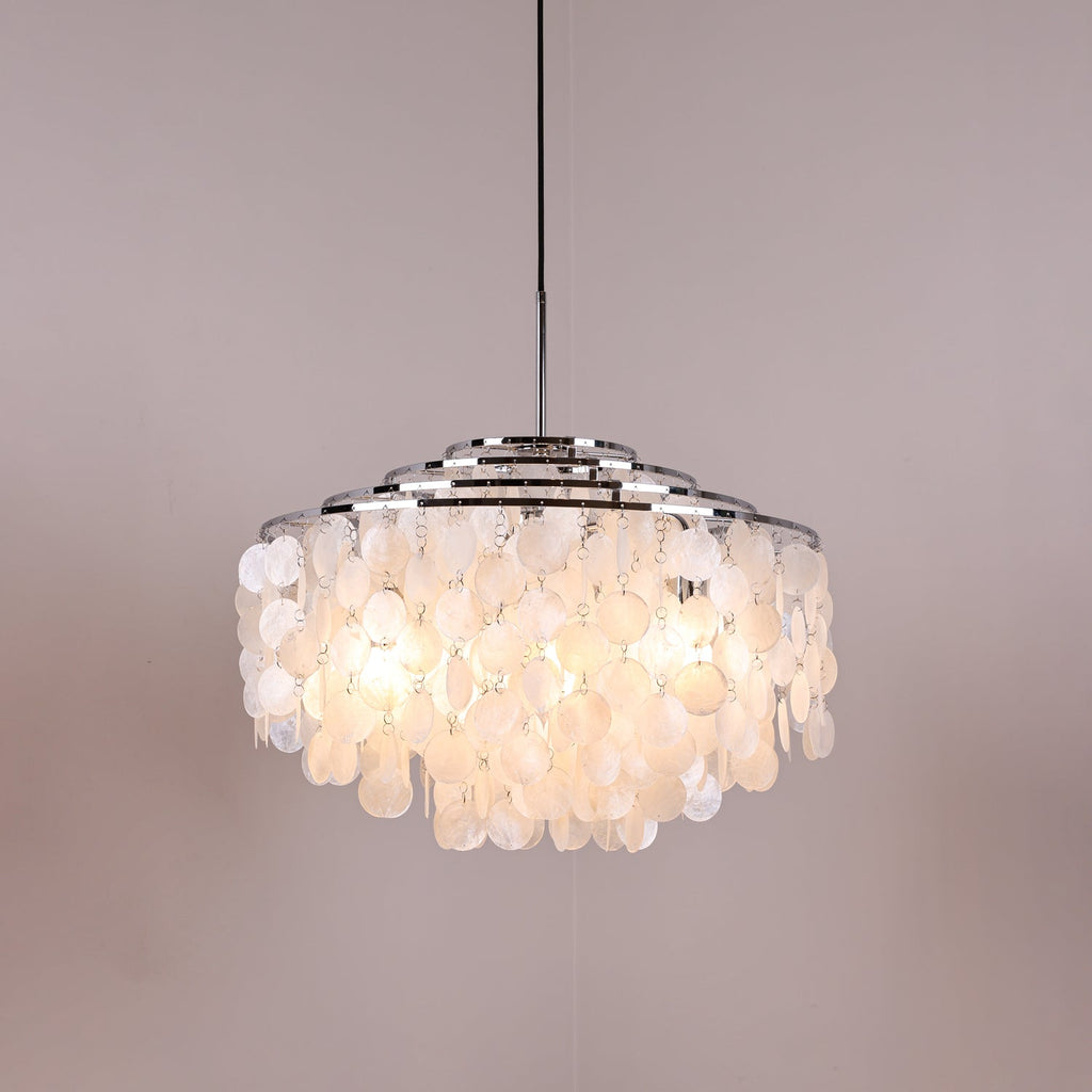 Lit Clérachic shell chandelier with chrome rings on a grey-pink backdrop, the discs glowing warm white from inside.