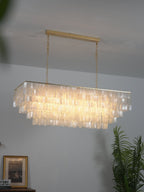 Lit rectangular Clérachic shell chandelier with a gold bar and chain, hung against a grey wall with a plant at one side.