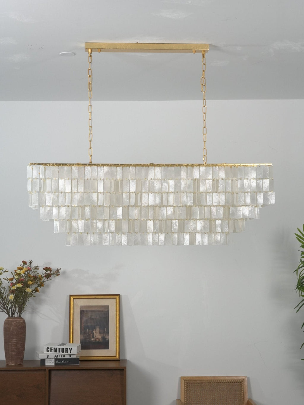 Unlit rectangular Clérachic shell chandelier with a gold bar and chains, its flat discs in rows, above a wooden sideboard.