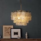 Lit Clérachic shell chandelier glowing warm against a dark blue-grey wall, with framed pictures on a wooden shelf below.