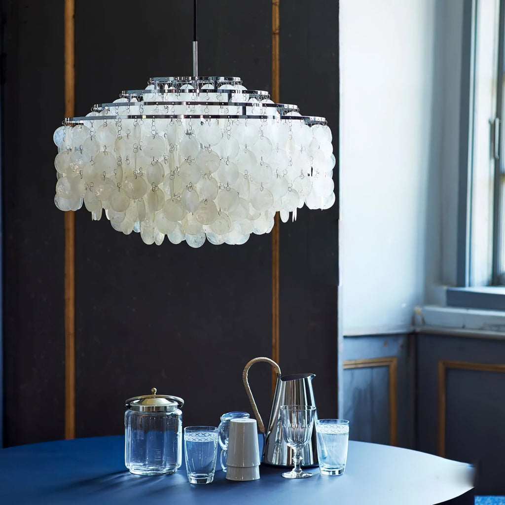 Chrome-ringed shell chandelier from Clérachic hanging above a navy table set with glass jugs, beside a tall window.