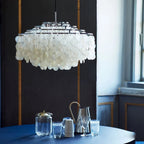 Chrome-ringed shell chandelier from Clérachic hanging above a navy table set with glass jugs, beside a tall window.
