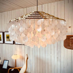 Gold-ringed Clérachic shell chandelier glowing in a plank-walled room, with framed pictures and a rattan shade behind it.