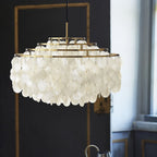 Unlit gold-ringed Clérachic shell chandelier hanging against a dark panelled wall with a window to one side.