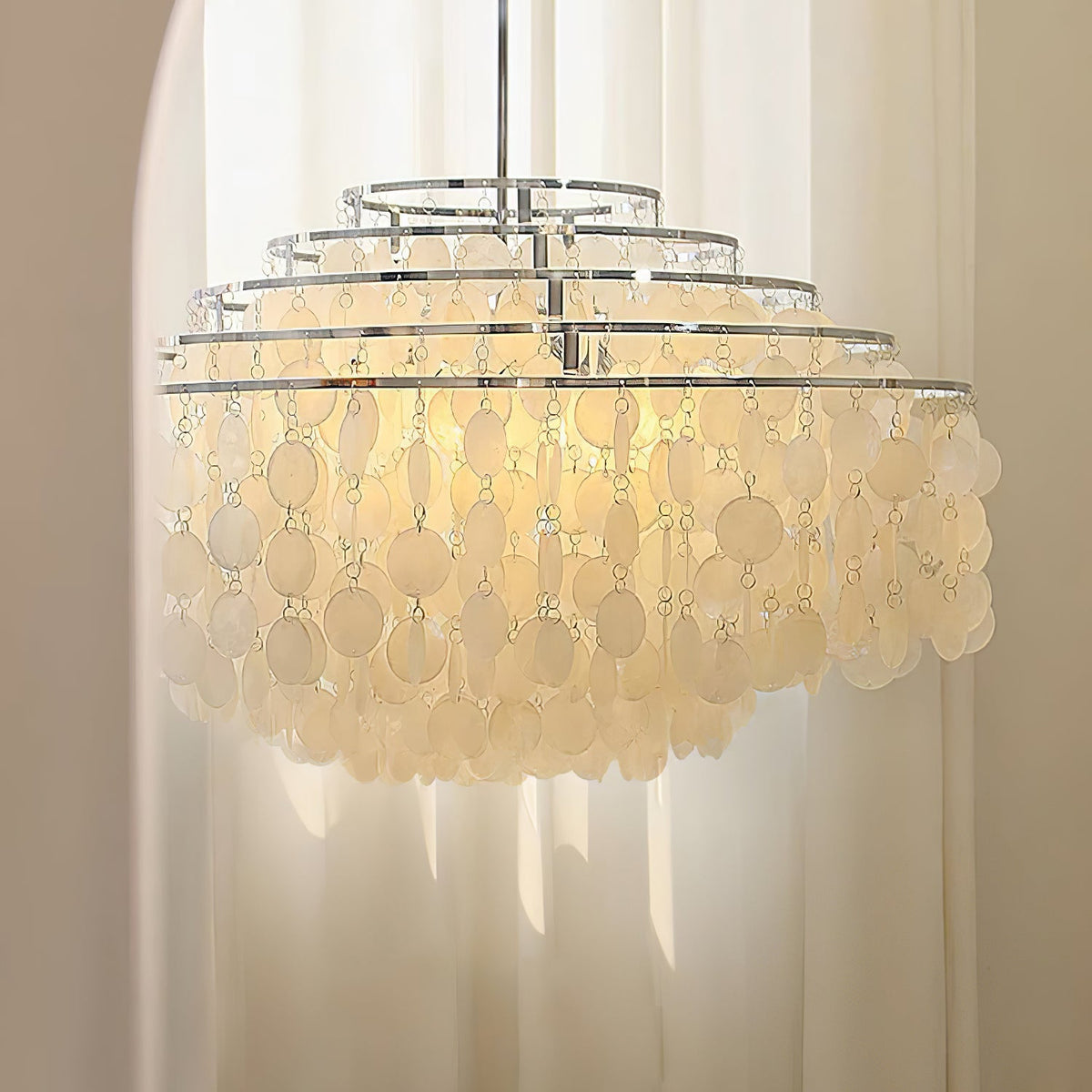 Chrome-ringed shell chandelier from Clérachic lit softly in front of cream curtains, its discs glowing warm white.