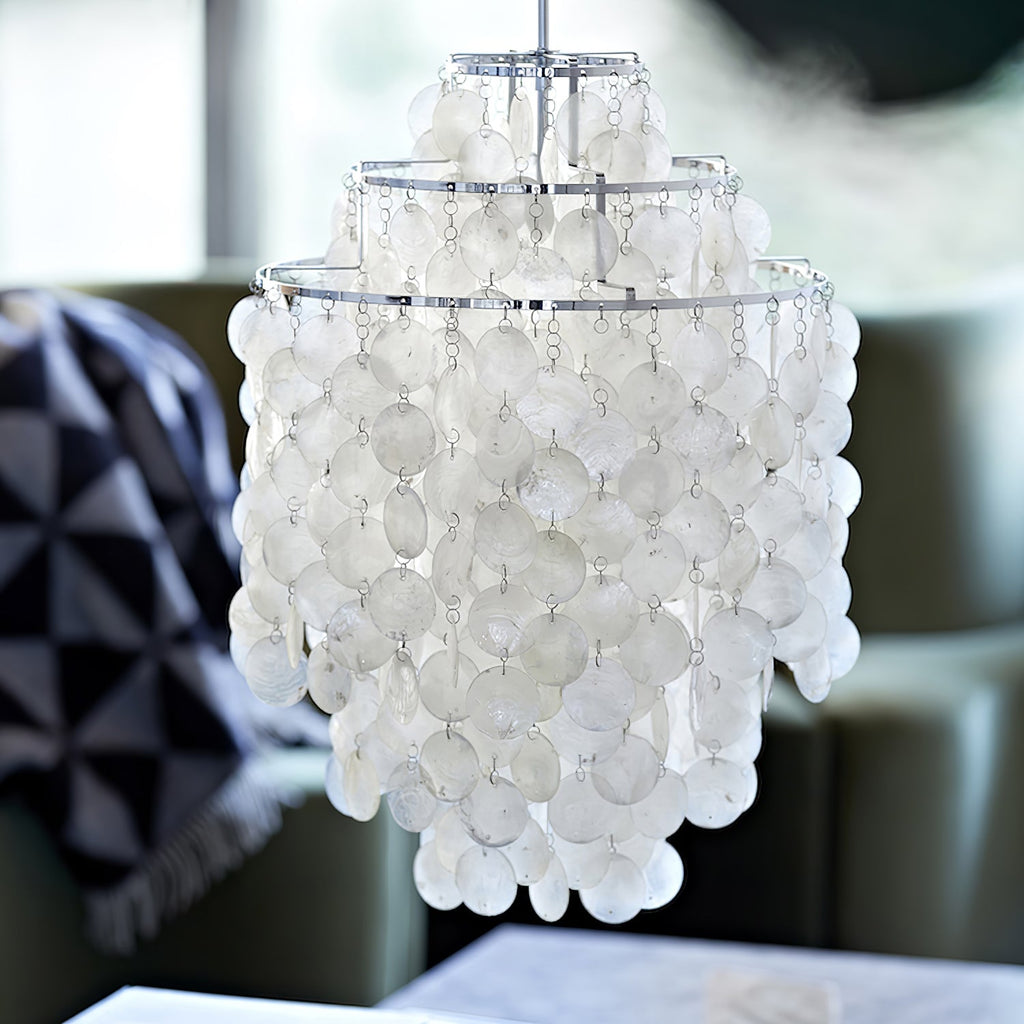 Close view of a chrome-ringed Clérachic shell chandelier, tiers of white discs in soft focus beside a grey armchair.