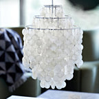 Close view of a chrome-ringed Clérachic shell chandelier, tiers of white discs in soft focus beside a grey armchair.