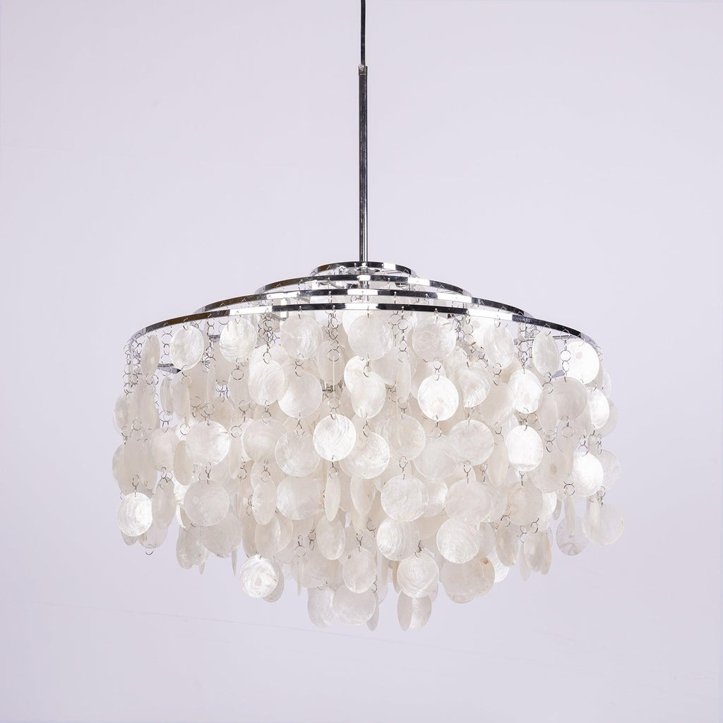 Chrome-ringed Clérachic shell chandelier on a pale lilac backdrop, its discs hanging in layers below a slim silver stem.
