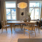 Gold-ringed Clérachic shell chandelier above an oval oak dining table and wishbone chairs, between two tall windows.