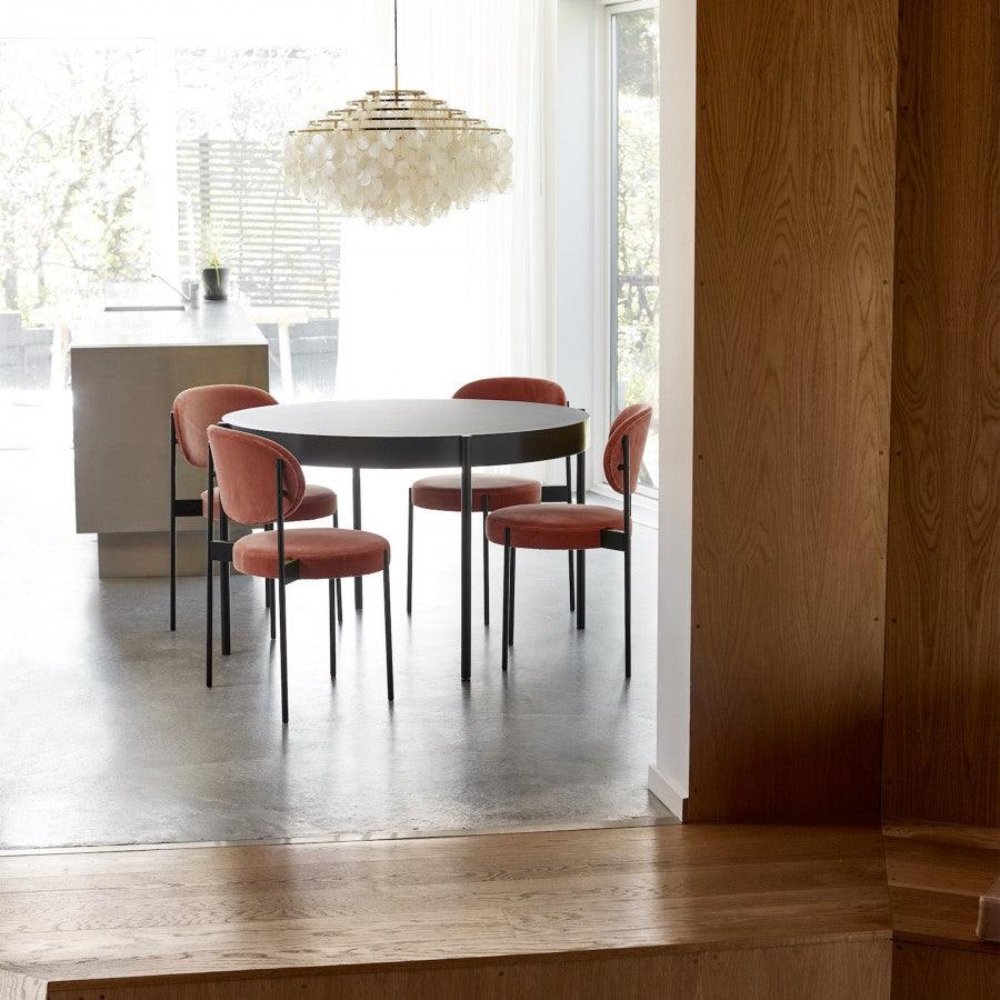 Distant view of a gold-ringed Clérachic shell chandelier above a round dining table with rust-red chairs by a large window.