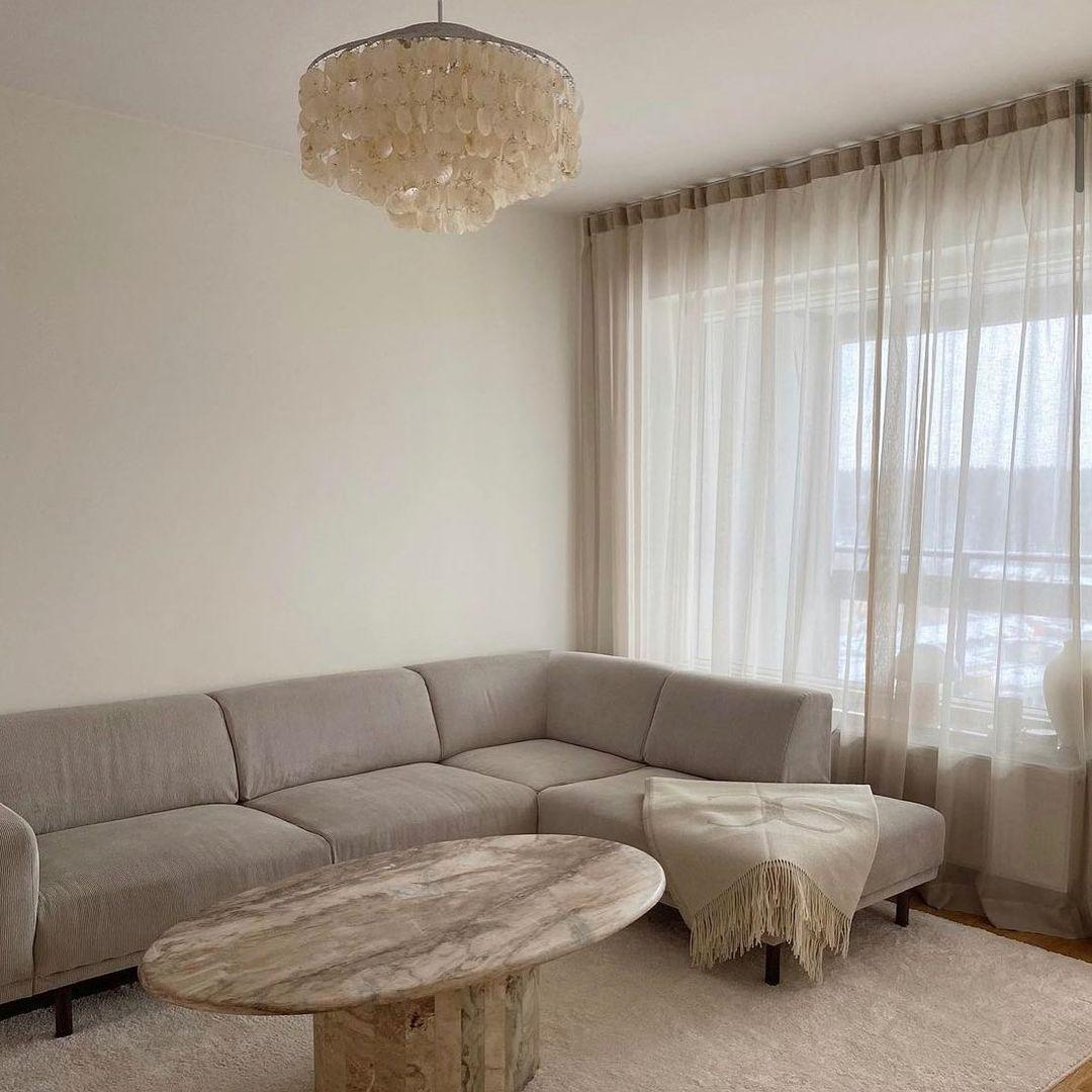 Clérachic shell chandelier hanging above a grey corner sofa and a marble-topped oval table, with sheer curtains at the window.