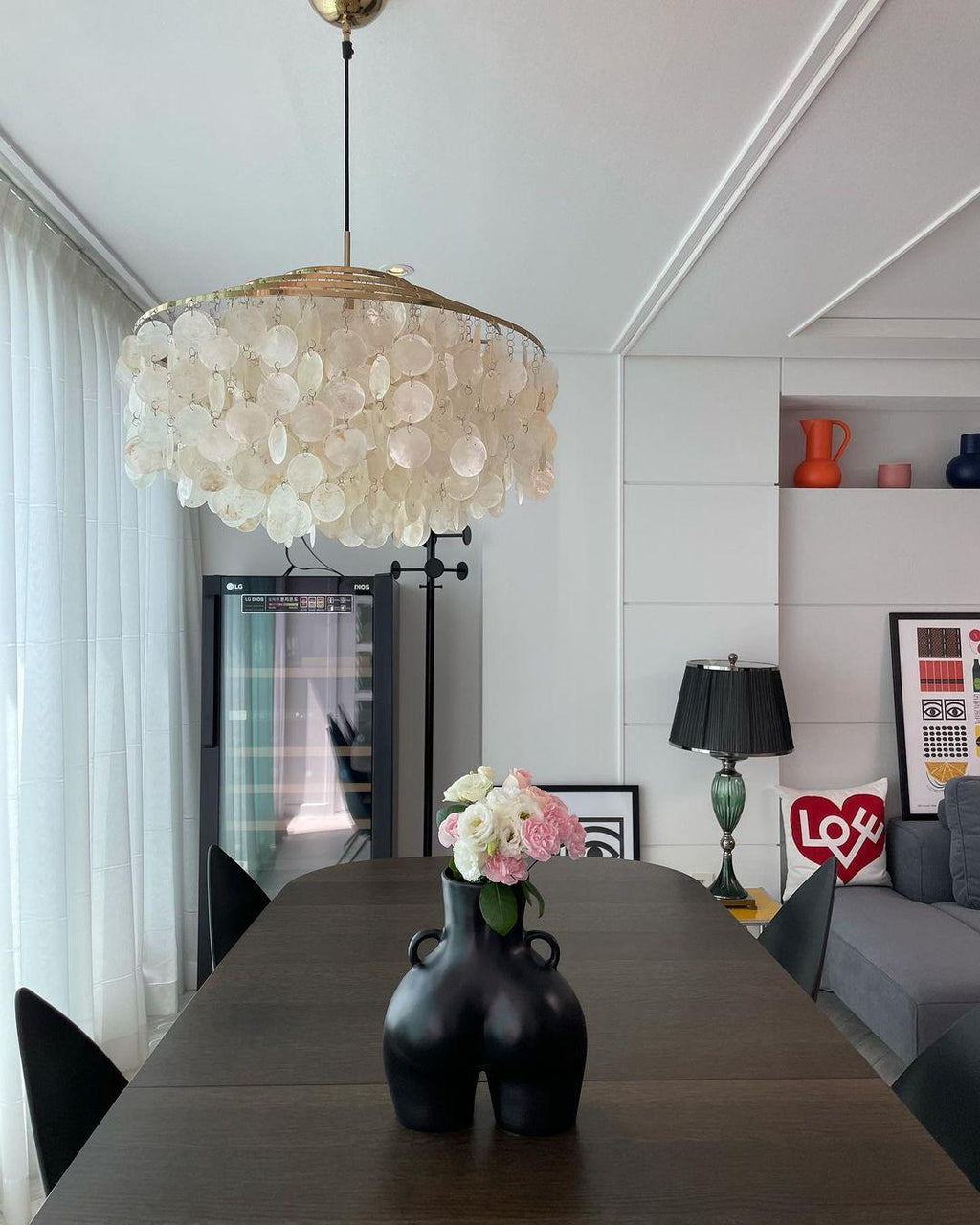 Gold-ringed Clérachic shell chandelier over a dark wood dining table with a black vase of pink flowers, in a bright room.