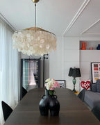 Gold-ringed Clérachic shell chandelier over a dark wood dining table with a black vase of pink flowers, in a bright room.