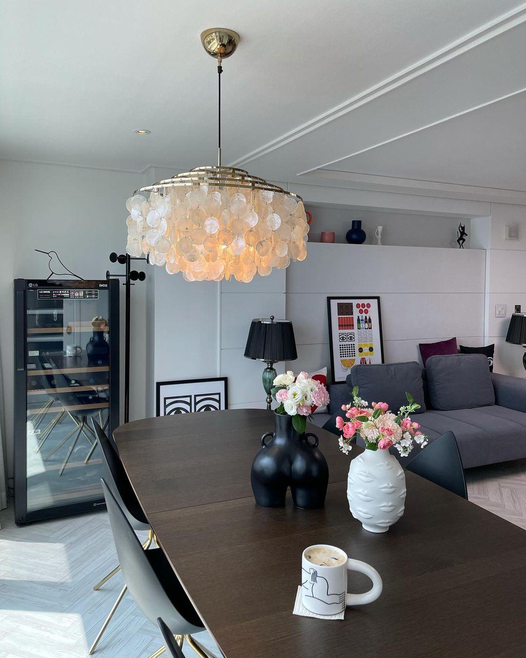 Lit Clérachic shell chandelier glowing amber above a dark dining table, with a black vase and a wine cabinet behind.