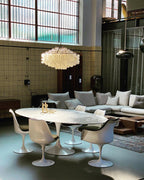 Clérachic shell chandelier over a white oval table and tulip chairs in a high-ceilinged room with a tiled wall and large windows.