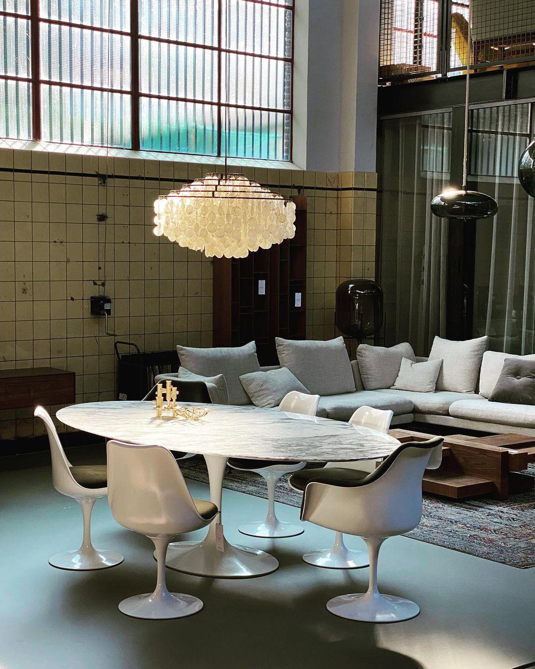 Clérachic shell chandelier over a white oval table and tulip chairs in a high-ceilinged room with a tiled wall and large windows.