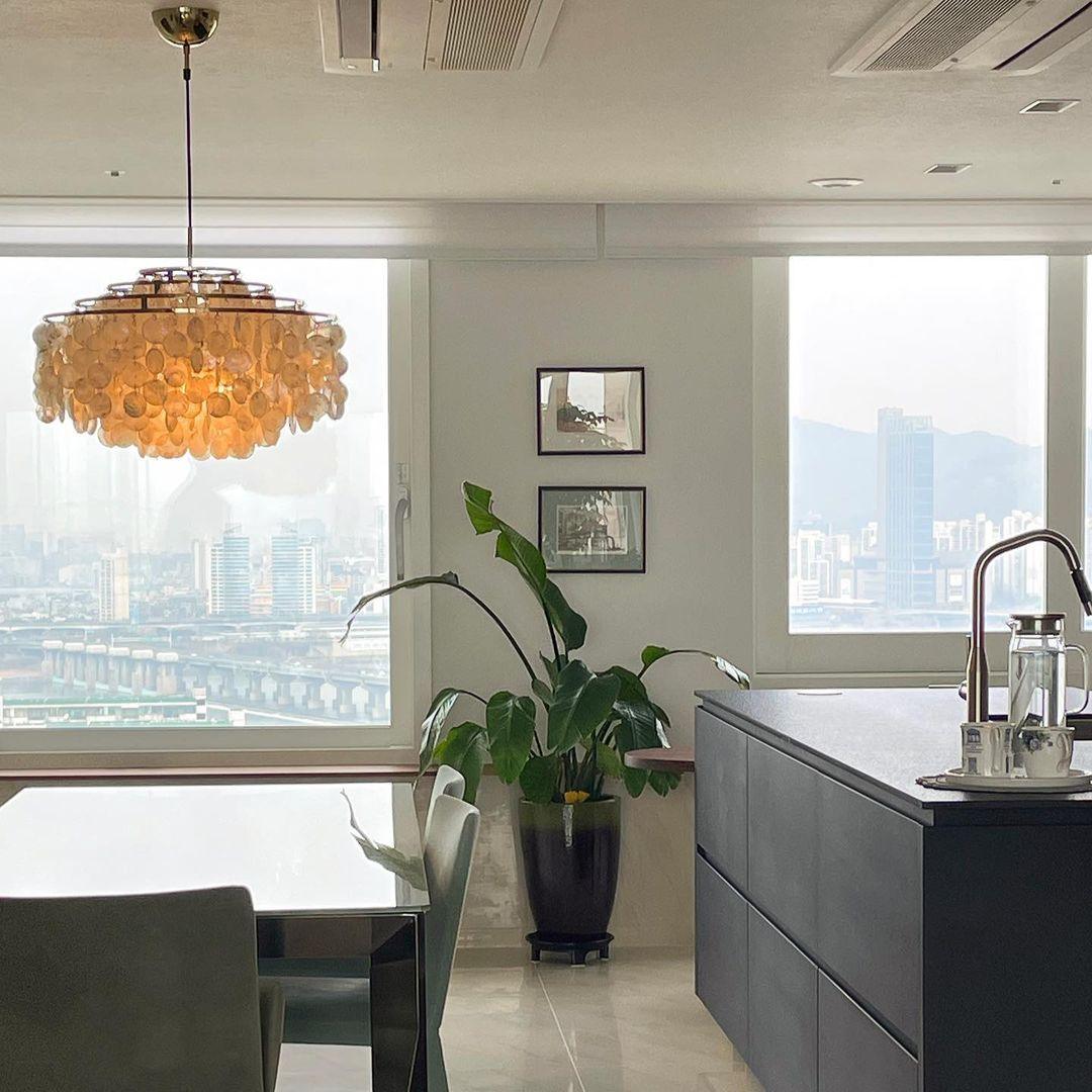 Clérachic shell chandelier glowing orange-gold in an apartment kitchen, with a city view through the windows behind.
