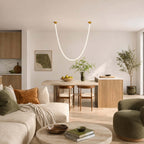 A single glowing U-shaped Clérachic pendant light hangs between gold plates over an open-plan dining and sofa area with an olive tree.