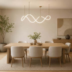 A Clérachic pendant with a looping glowing strip hangs above a long oak table and cream chairs, in a warm room with an olive tree.