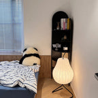 Clérachic pear-shaped silk floor lamp, lit in a bedroom corner beside a black arched bookshelf and striped bed linen.