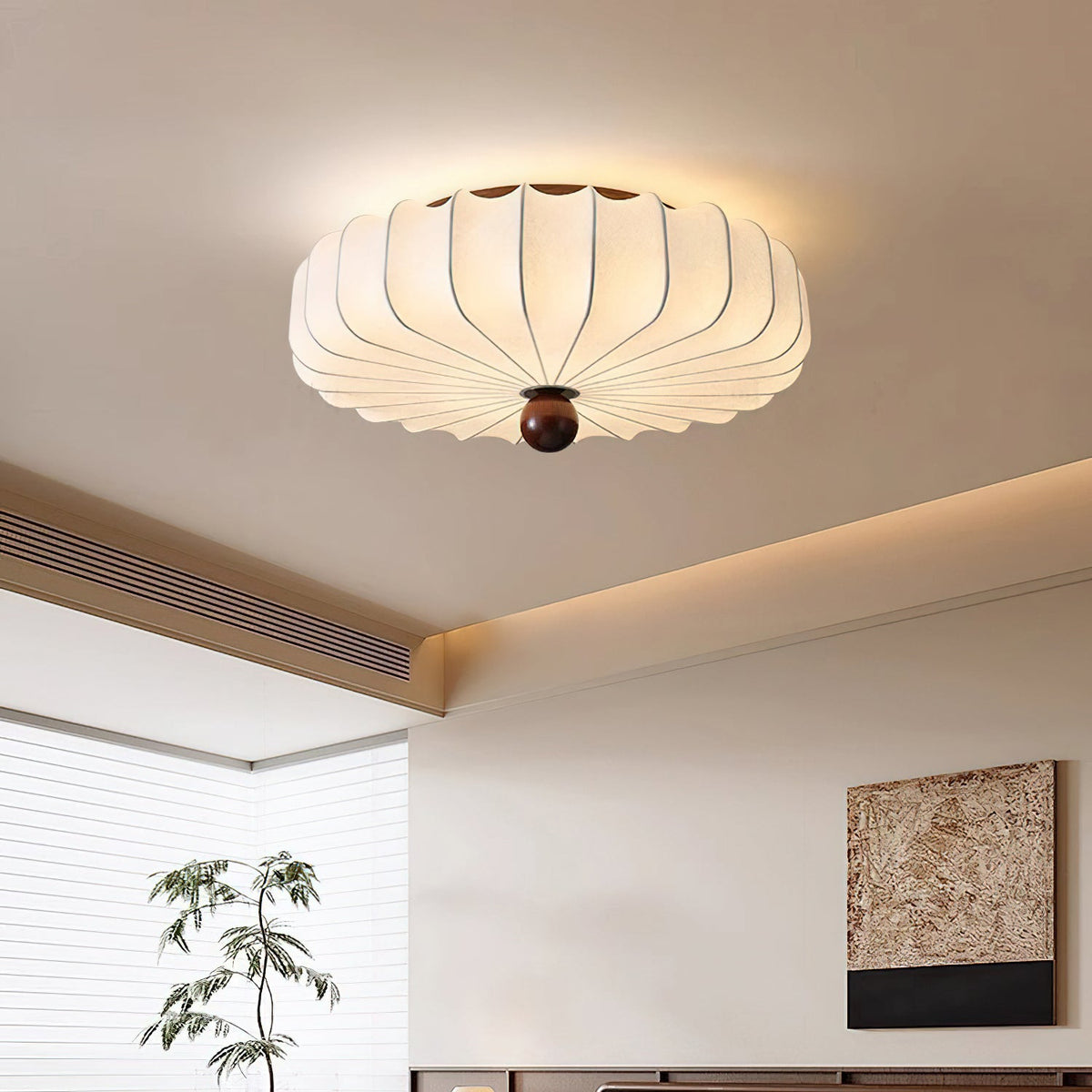A lit Clérachic flush-mount ceiling light in a bedroom with a cove-lit ceiling, a tall plant and a textured wall panel.