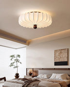 Looking up at a lit Clérachic ceiling light over a bed with layered bedding, a bedside lamp and a potted tree by the window.