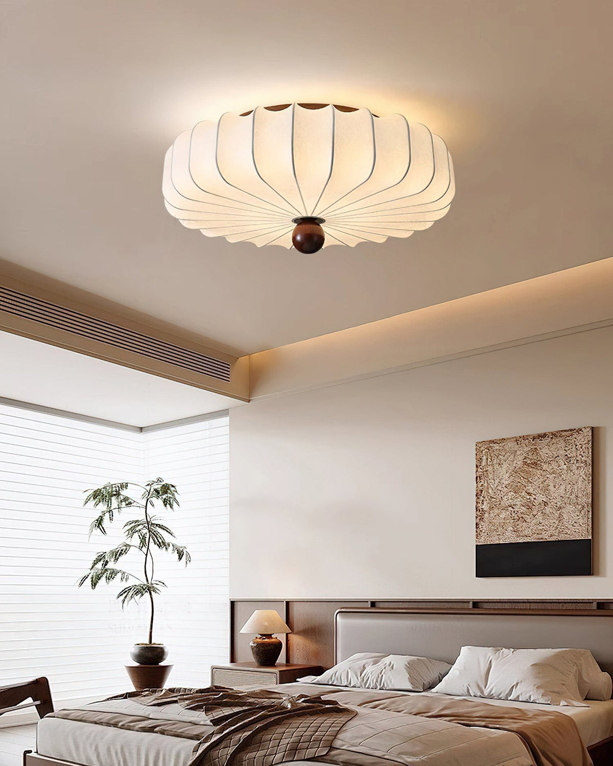 Looking up at a lit Clérachic ceiling light over a bed with layered bedding, a bedside lamp and a potted tree by the window.